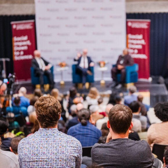 An audience watches a Stone Center panel discussion on stage. Three panelists sit in chairs facing the audience, with two large banners and a screen displaying the topic 'Conversations on Inequality and Public Policy' in the background.